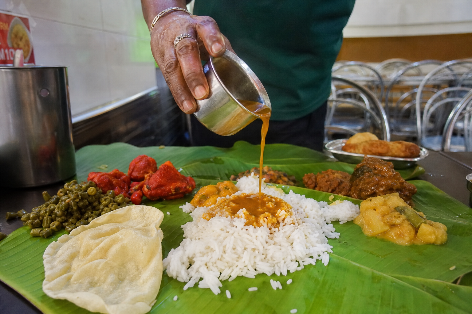 Close-up eye-level shot of a banana leaf rice spread at a Brickfields restaurant in Kuala Lumpur with rice, curries, papadom, and rasam being poured
